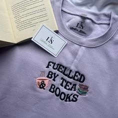 an open book sitting on top of a table next to a t - shirt that says fueled by tea and books