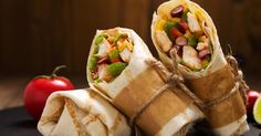 two burritos wrapped in brown paper with vegetables and meat on top, next to sliced tomatoes