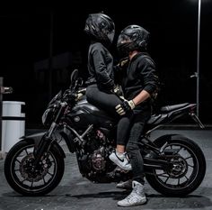 two people are sitting on a motorcycle in the dark with their helmets on and one person is wearing black