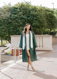 a woman in a white dress and green coat is standing on a brick walkway with trees behind her