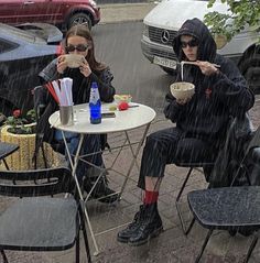 two people sitting at a table in the rain eating food and drinking from paper cups