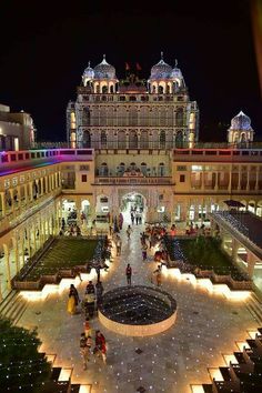 people are walking around in front of an ornate building at night with lights on the ground