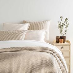 a bed with white sheets and pillows in a room next to a nightstand, potted plant