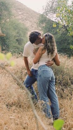 a man and woman are kissing in the tall grass with trees behind them on a hill