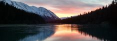 a lake surrounded by trees and mountains with the sun setting in the sky above it