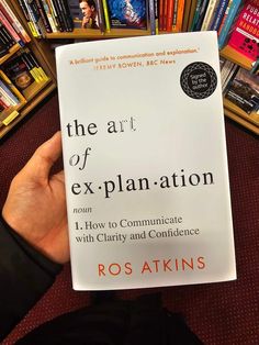 the art of explanation by ros akins is being held up in front of bookshelves