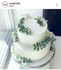 a white wedding cake decorated with greenery and flowers on a table next to a window