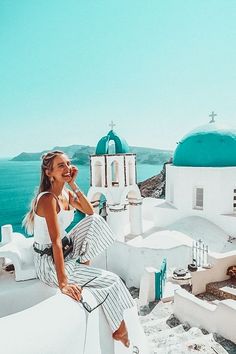 a woman sitting on top of a white building next to the ocean with blue domes