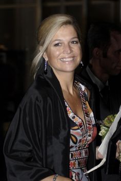 a woman in a black jacket holding a bouquet and smiling at the camera with other people behind her