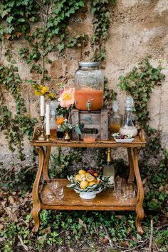 a table topped with fruit and drinks next to a wall covered in vines, flowers and greenery