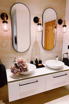 A modern bathroom featuring dual oval mirrors framed with black edges, highlighted by brass and glass globe wall lights. The warm glow complements the neutral beige tile backdrop, white countertops with dual sinks, fresh hydrangea flowers, and minimalist black bathroom accessories. Bathroom Lighting Inspiration, Bathroom Sanctuary, Globe Wall Light, Minimalist Accessories, Bathroom Goals, Mirror Design, Elegant Bathroom, Interior Stylist, Lighting Inspiration