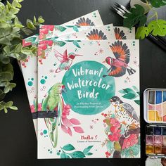 three watercolor books are sitting on a table next to some paintbrushes and flowers