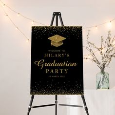 a black and gold graduation party welcome sign on an easel with lights in the background