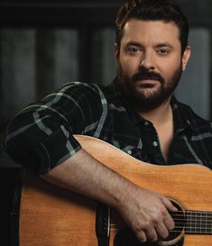 a bearded man holding an acoustic guitar in his right hand and looking at the camera