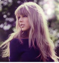 a woman with long blonde hair standing in front of trees and looking off into the distance