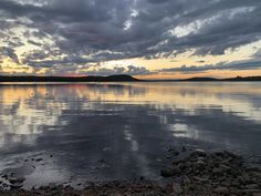 the sky is reflected in the calm water at sunset, with rocks and pebbles on the shore