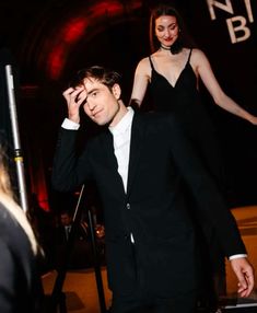 a man in a tuxedo walking down a red carpeted hallway with two women behind him