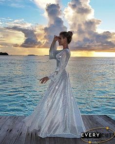 a woman in a long white dress standing on a dock near the ocean with clouds