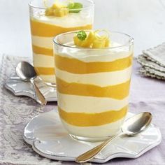 two glasses filled with dessert sitting on top of a white and yellow table cloth next to silver spoons