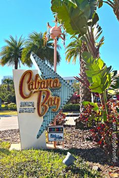 a sign that is in the grass near some trees and bushes with palm trees behind it