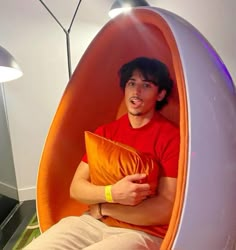 a man is sitting in an orange and white egg shaped chair holding a yellow pillow