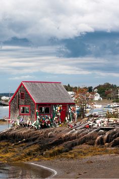 a small red house sitting on top of a rocky shore next to the ocean with boats in the water