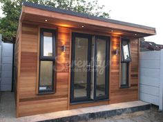 a small wooden building with glass doors and lights on the outside, next to a fence