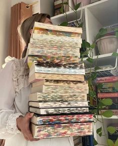 a woman holding a stack of books in front of her face while standing next to a plant