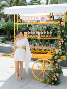 a woman standing in front of a yellow cart with bottles on it and pink flowers