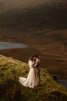 a man and woman standing on top of a grass covered hill next to a lake