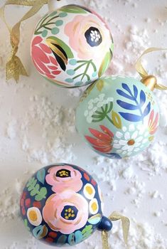 three ornaments with flowers painted on them and gold ribbons hanging from the top, along with white snow