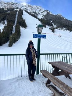 a woman standing next to a wooden bench on top of a snow covered slope in front of a mountain