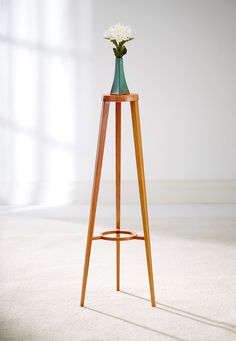 a tall wooden stand with a vase on top and flowers in the middle, against a white wall