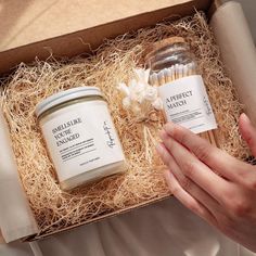 someone is holding two jars of candles in a box with straw and flowers on it