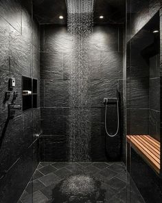 a shower head in the middle of a black tiled bathroom with wooden bench under it