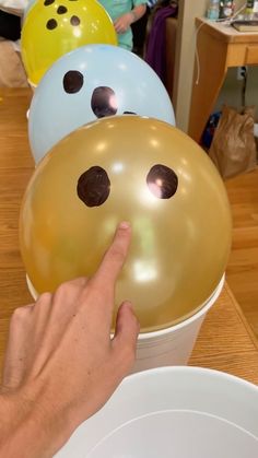 a hand pointing at several balloons in a bucket on a table with other people around