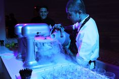 a man pouring drinks into glasses in front of an ice machine on a table with people standing around