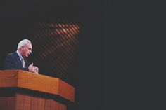 a man standing at a podium with his hands folded in front of him and looking off into the distance