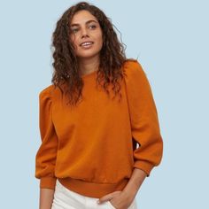 a woman with curly hair wearing an orange sweater and white pants, standing in front of a blue background