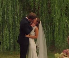 a bride and groom kissing in front of a willow tree during their wedding ceremony at the park