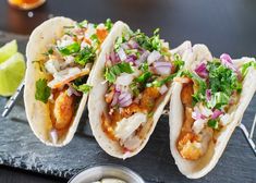 three fish tacos with onions, cilantro and lime on a slate board