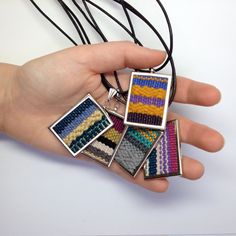 a person's hand holding four square pendants in different colors and patterns on black cord