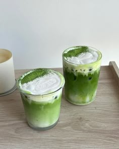 two glasses filled with green drinks sitting on top of a table next to a cup