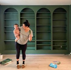 a woman holding a baby while standing in front of bookshelves with other items on the floor