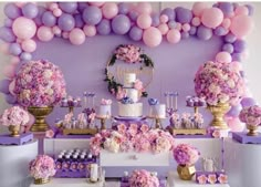 a table topped with lots of pink and purple flowers next to a wall covered in balloons