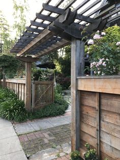 a wooden pergolated area with plants and flowers on the side walk in front of it