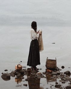 a woman is standing on the rocks by the water looking at her purse and clothes