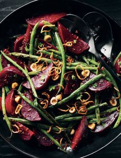 a black plate topped with green beans and beets next to silver spoons on a wooden table