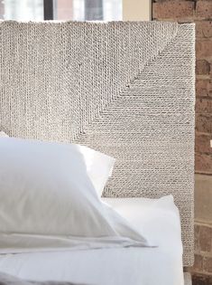 a bed with white sheets and pillows in front of a brick wall that has a woven headboard