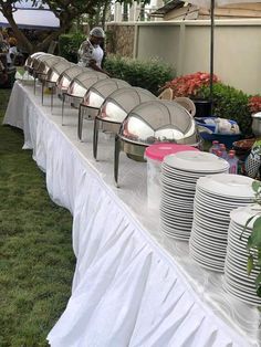 there are many plates and cups on the long table with white cloth draped over it